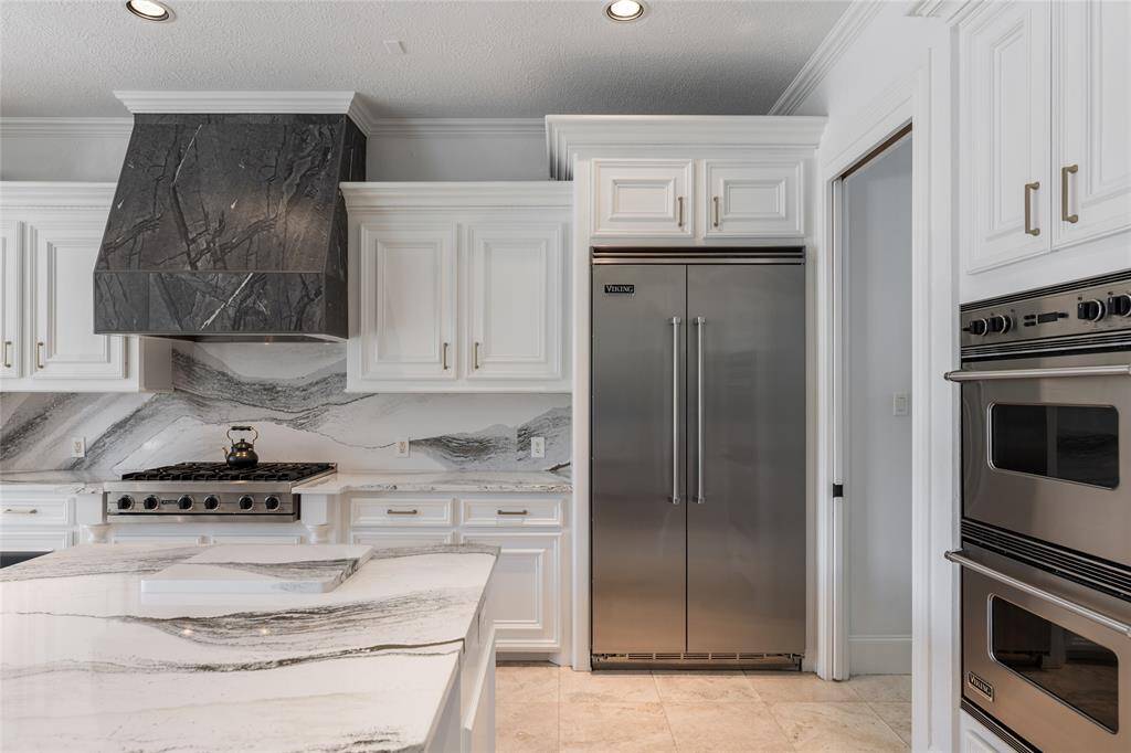 This modern kitchen features elegant white cabinetry, a striking quartz backsplash and countertops, and high-end stainless steel appliances, including a double-door refrigerator and a built-in oven. The spacious design is complemented by a large island, perfect for entertaining.