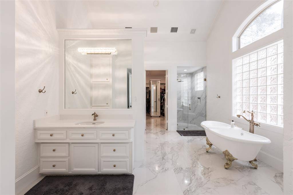 This bathroom features elegant marble flooring, a classic freestanding bathtub, and a spacious glass-enclosed shower. The room is bright with a large window, and the vanity includes ample storage and a stylish mirror.