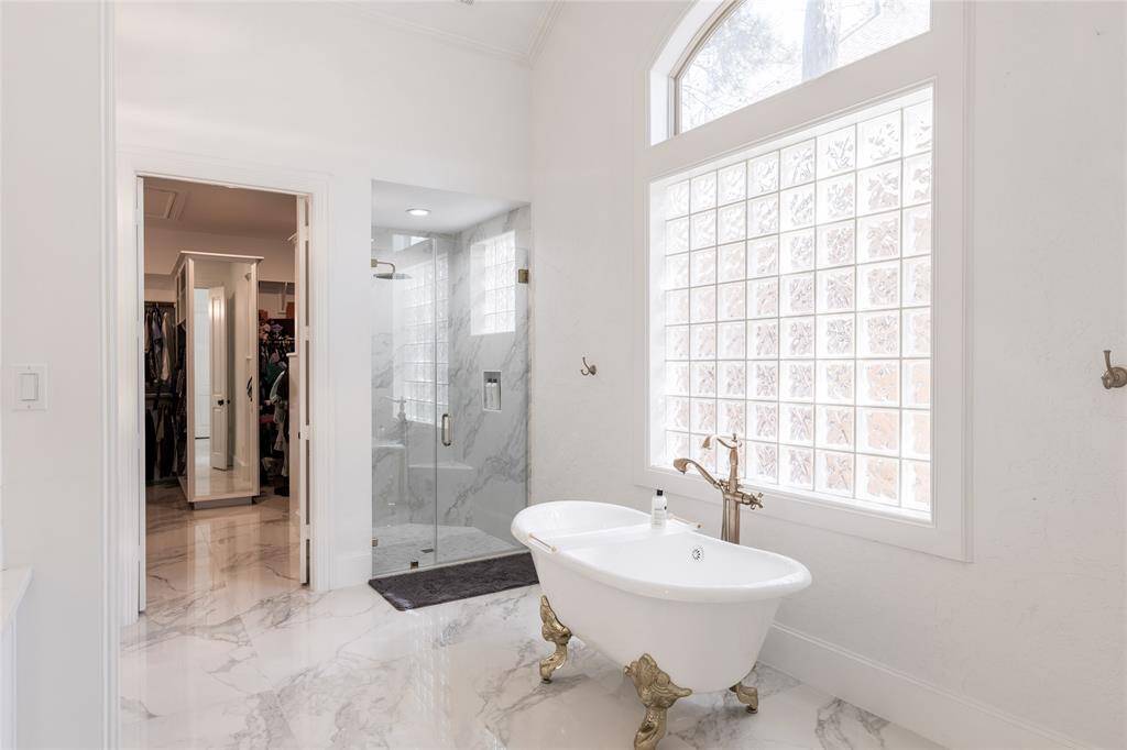 This elegant bathroom features a freestanding clawfoot tub with ornate fixtures, a spacious glass-enclosed shower, and a large window with frosted glass blocks for natural light. It leads into a walk-in closet with ample storage and a full-length mirror. The marble flooring adds a touch of luxury.