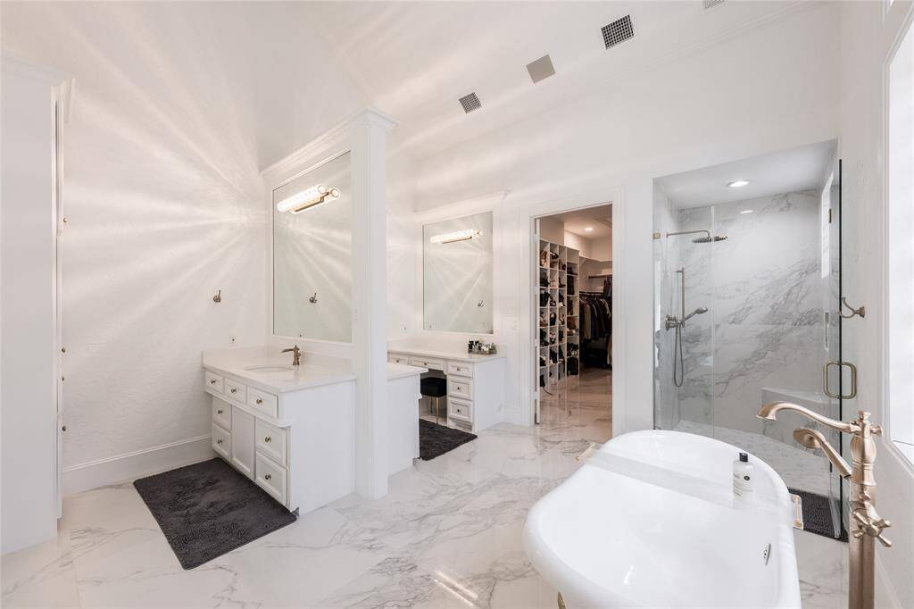 This spacious bathroom features elegant marble flooring, a freestanding bathtub, and a glass-enclosed shower. Dual vanities offer ample storage, and the room is connected to a large walk-in closet, enhancing convenience and luxury.