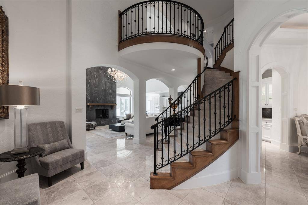 This elegant home features a grand entrance with a curved staircase and wrought iron railing. High ceilings and a chandelier add sophistication, while archways lead to a spacious living area with a modern fireplace. The neutral palette and polished floors create an inviting ambiance.