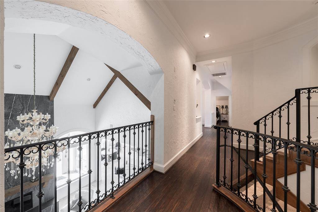 This elegant home features a hallway with hardwood floors, leading to a curved staircase with iron railings. An open archway reveals a vaulted ceiling with exposed wooden beams and a luxurious chandelier, adding a touch of sophistication.
