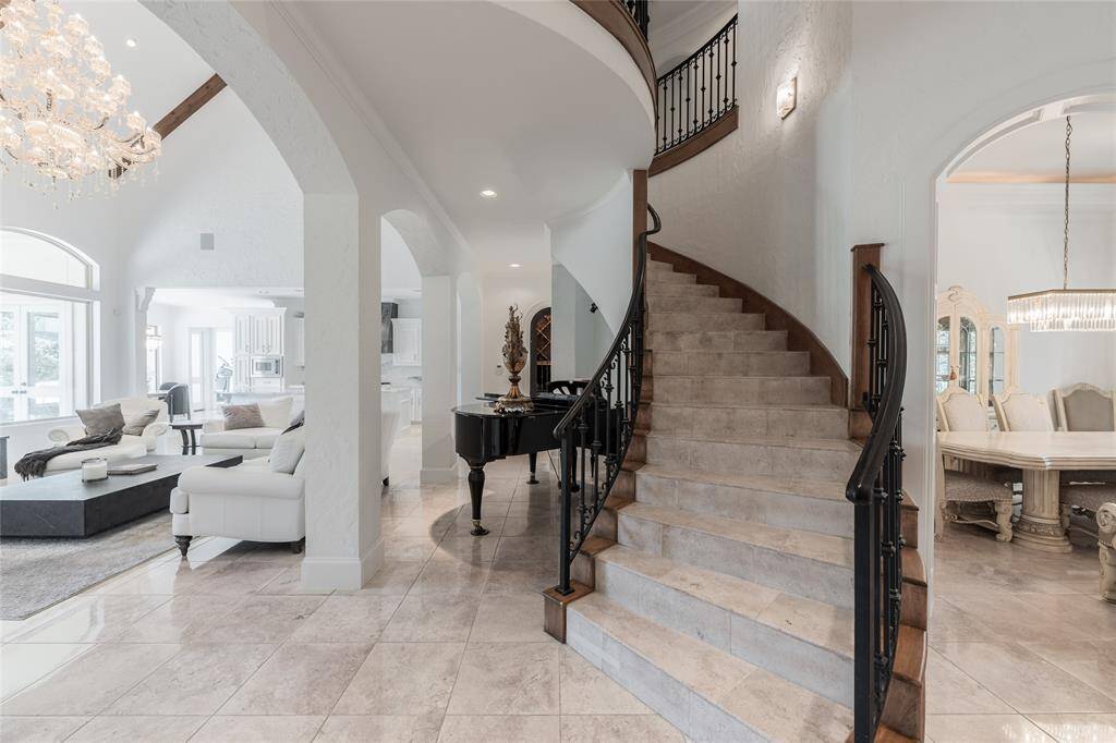 This elegant home features a grand, curved staircase and open-concept living spaces. The entrance highlights a polished travertine stone floor and a sophisticated interior with a chandelier, and spacious seating areas. Natural light floods the rooms, enhancing the airy and luxurious ambiance.
