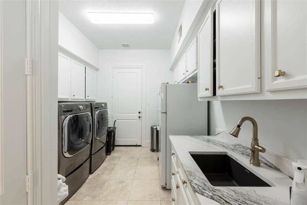 This is a modern laundry room featuring a sleek design, ample white cabinetry for storage, and a quartz countertop with a stylish sink. The space is well-lit and has a tiled floor, providing a clean and functional area for laundry tasks.