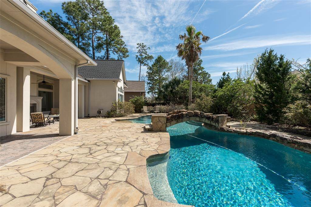 This photo showcases a spacious backyard featuring a beautiful, curvy swimming pool surrounded by a stone patio. The area is lined with lush greenery and trees, offering a serene and private outdoor space. The house exterior is modern, with covered seating areas perfect for relaxation or entertaining.