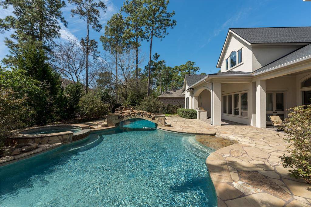 This photo showcases a beautiful backyard with a spacious swimming pool, hot tub, and stone patio area. The house features large windows and a covered patio, surrounded by lush greenery and tall trees, offering a serene and private outdoor space.