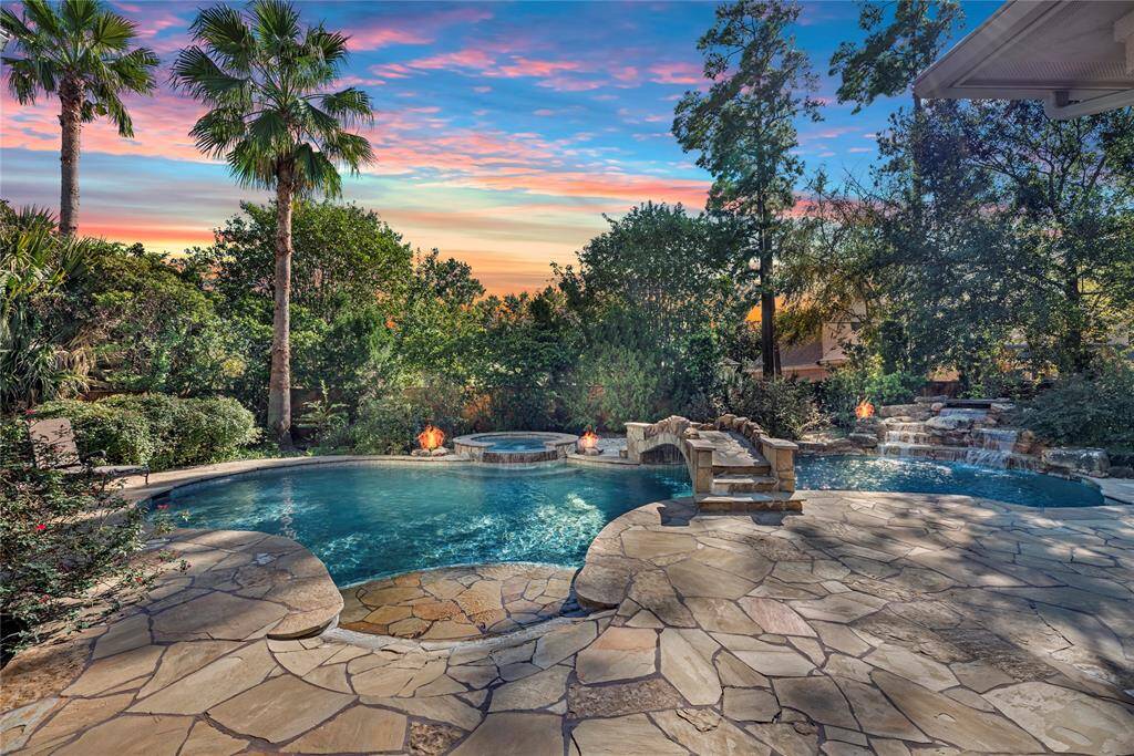 This photo showcases a luxurious backyard featuring a sprawling pool with a stone bridge, surrounded by lush landscaping and tall palm trees. There's a cozy fire feature and a waterfall, all under a stunning sunset sky, creating a serene and inviting outdoor oasis.