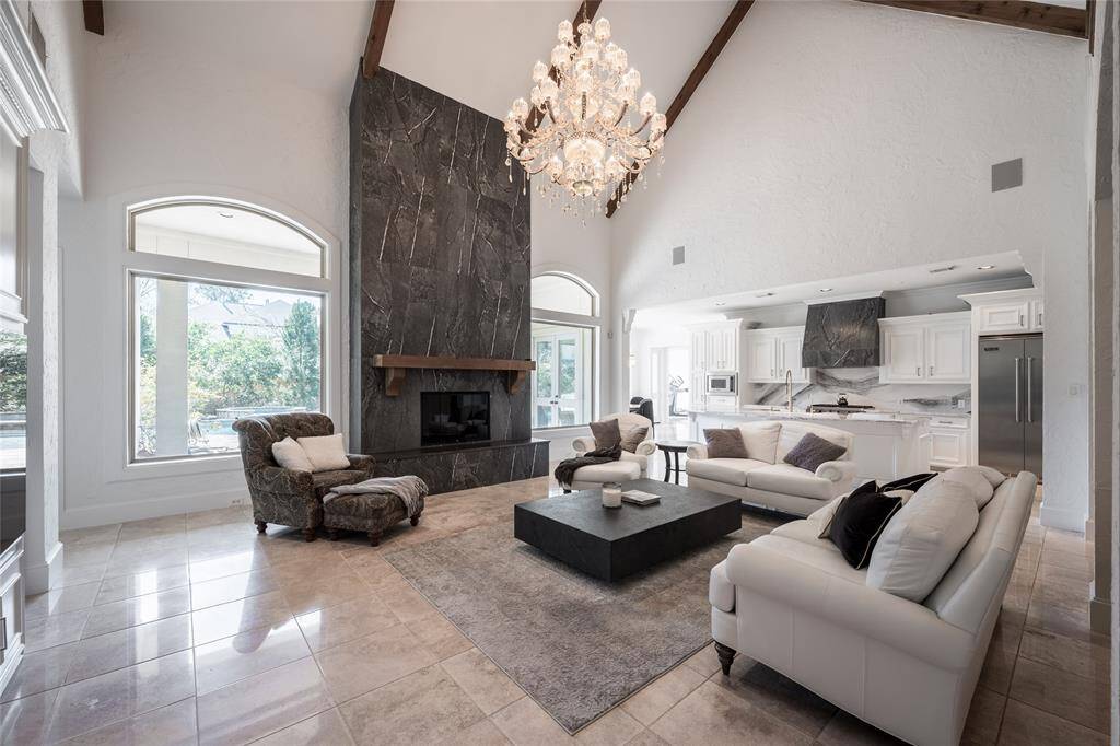 This spacious living area features high vaulted ceilings, a stunning chandelier, and a dramatic black porcelain fireplace. Large windows bring in natural light, complementing the elegant white kitchen with modern appliances. The open layout includes stylish seating and a sophisticated, neutral color palette.