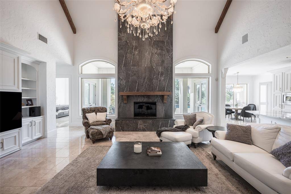 This elegant living room features a high ceiling with exposed beams and a stunning chandelier. The focal point is a dramatic porcelain fireplace flanked by large windows, allowing natural light to flood the space. The open layout connects seamlessly to the dining area, creating a spacious and inviting atmosphere.
