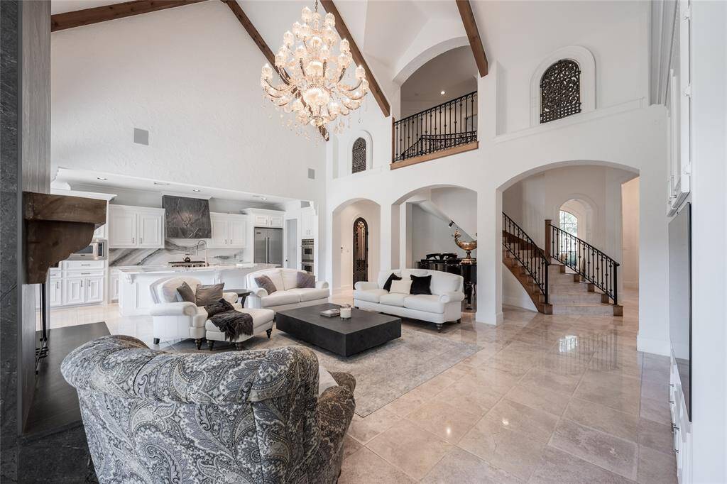 This elegant living space features high ceilings with exposed wooden beams, a grand chandelier, and an open floor plan. The room includes a cozy seating area with modern furnishings, a sleek kitchen with white cabinetry, and a beautiful staircase with wrought iron railings. Natural light floods the space, enhancing its luxurious and spacious feel.
