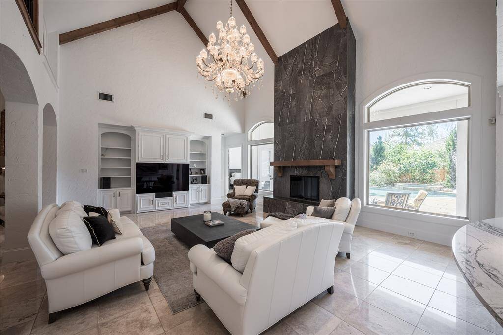 This spacious living room features high vaulted ceilings with wooden beams, a grand chandelier, and a large porcelain fireplace. The room is brightened by large arched windows offering a view of the backyard.