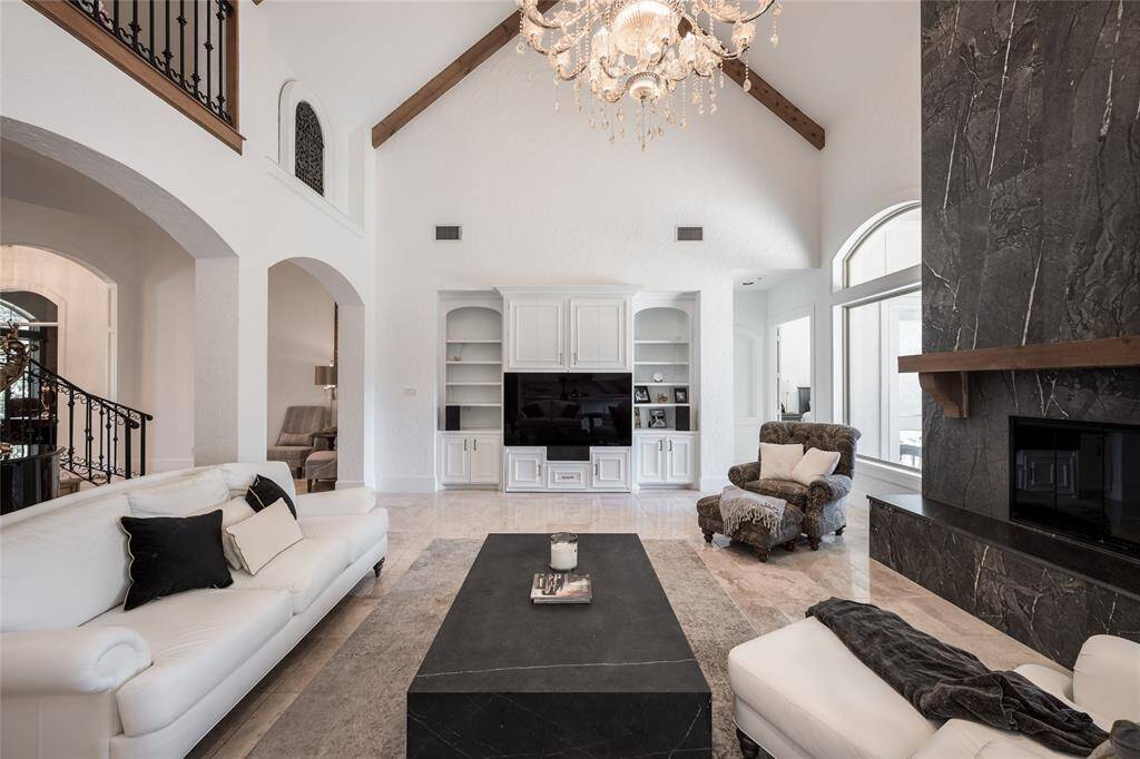 This spacious living room features high vaulted ceilings with exposed wooden beams, a grand chandelier, and a stylish porcelain fireplace. It includes a large built-in entertainment center, and ample natural light from large arched windows, creating a luxurious and inviting atmosphere.