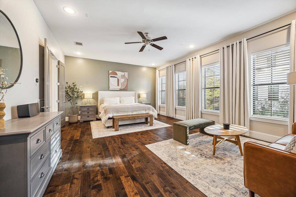 Expansive primary suite includes space for a king bed and a cozy sitting area, surrounded by a wall of windows and custom window treatments. Hardwood flooring and neutral tones create a relaxing retreat.