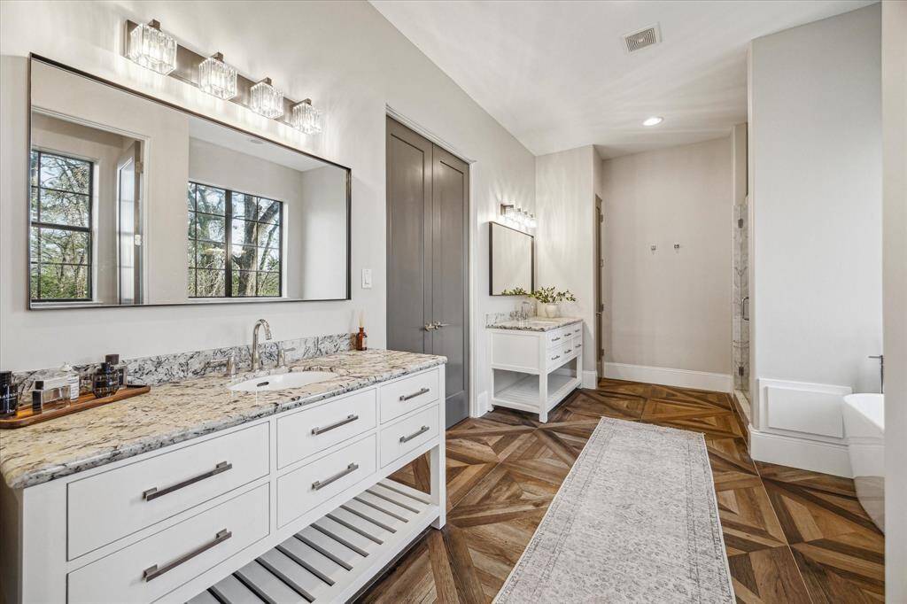 Luxurious primary bathroom offers dual vanities with granite counters, framed mirrors, and designer lighting. Beautiful wood-patterned tile flooring adds a touch of sophistication.