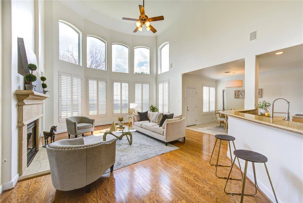 A soaring two-story living room filled with natural light from expansive windows. Gleaming wood floors flow throughout, complemented by a cozy fireplace and an open-concept design that seamlessly connects to the kitchen.