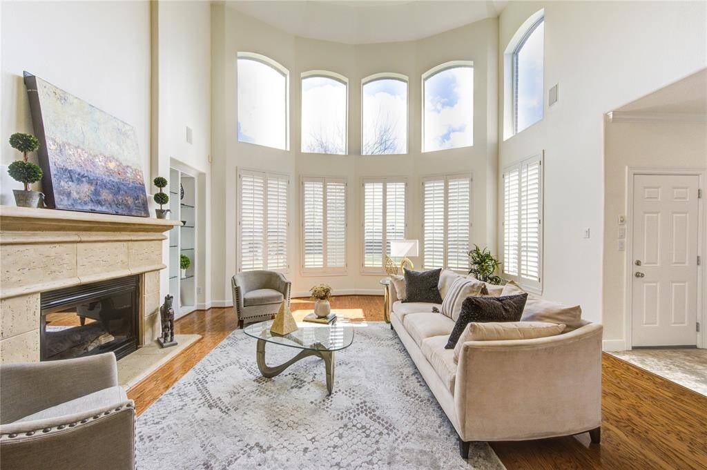 Step into a breathtaking two-story living room filled with natural light from a wall of windows, showcasing the home’s freshly painted interior and elegant wood floors. A cozy gas fireplace with a natural stone mantel serves as a stunning focal point