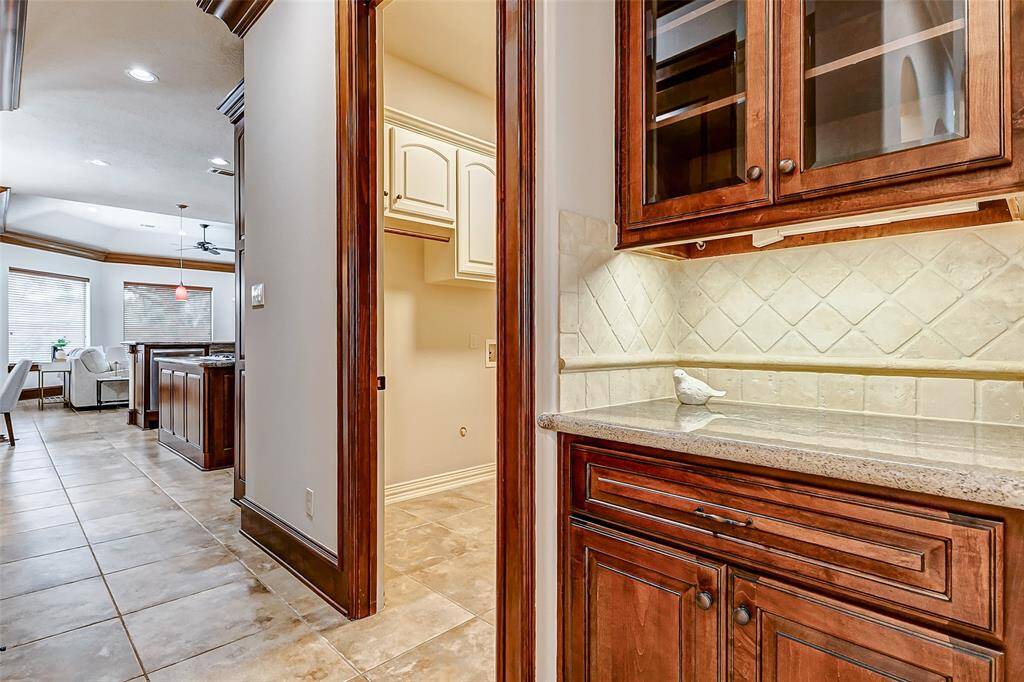 Beautifully designed butler’s pantry featuring rich wood cabinetry with glass-front uppers, granite countertops, and a stylish tumbled stone backsplash. Under-cabinet lighting and electric plugs enhances the space, providing both functionality and ambiance. This space offers additional storage and prep space, perfect for entertaining.