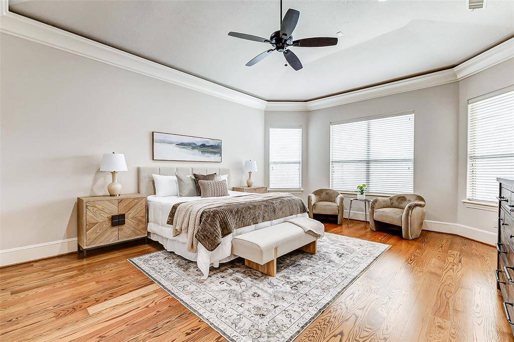 Spacious and elegant, this first-floor primary bedroom features high tray ceilings with crown molding, beautiful hardwood flooring, and large windows that fill the room with natural light. The generously sized layout offers ample space for a sitting area, creating a serene retreat. The room is equipped with a ceiling fan and recessed lighting for added comfort.