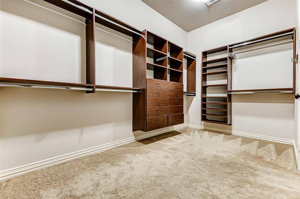 This primary walk-in closet is designed for both functionality and luxury, featuring Spaceman custom built-ins that optimize organization. The layout includes double hanging rods, open shelving, built-in drawers, and dedicated shoe storage to accommodate an extensive wardrobe. Rich wood finishes enhance the space, while plush carpeting provides comfort underfoot. Conveniently connected to the primary bath, this closet blends practicality with high-end design for effortless daily living.