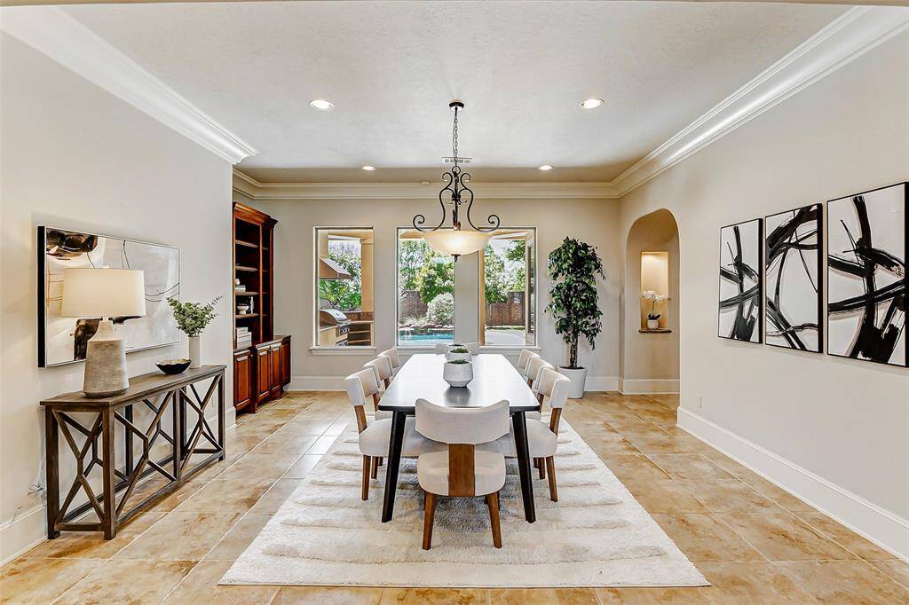 Elegant formal dining room featuring 12-inch crown molding, recessed lighting, and an iron-accented chandelier that adds a touch of sophistication. Built-ins with shelving provides ample storage and display space. Large windows fill the space with natural light and offer a seamless view of the backyard and pool/spa. The open layout allows for effortless entertaining, making this space both functional and inviting. (Can also be used as the formal living area).