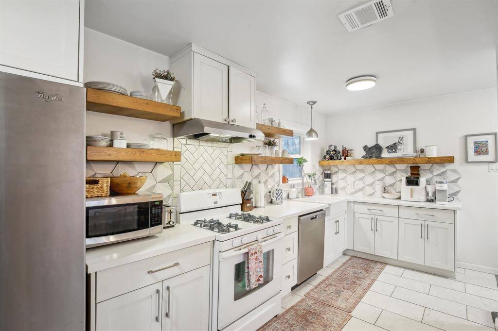 This kitchen features a modern design with white cabinetry and quartz countertops, complemented by wooden open shelves. It includes a gas stove, stainless steel appliances, and a stylish tile backsplash.