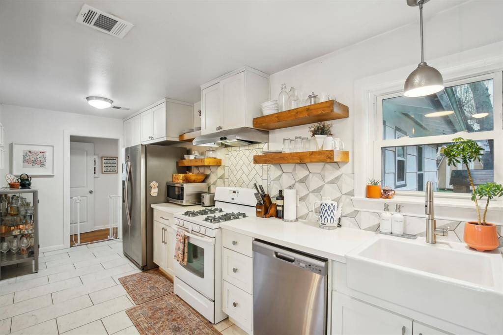 This bright kitchen features modern white cabinetry, stainless steel appliances, and open wooden shelves. The geometric tile backsplash adds a stylish touch, while a large sink and ample counter space enhance functionality.