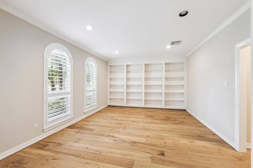 The home office/study features double arched windows with custom coastal shutters overlooking the front lawn, updated engineered wood floor, crown molding, recessed lighting and a wall of built-in shelving.