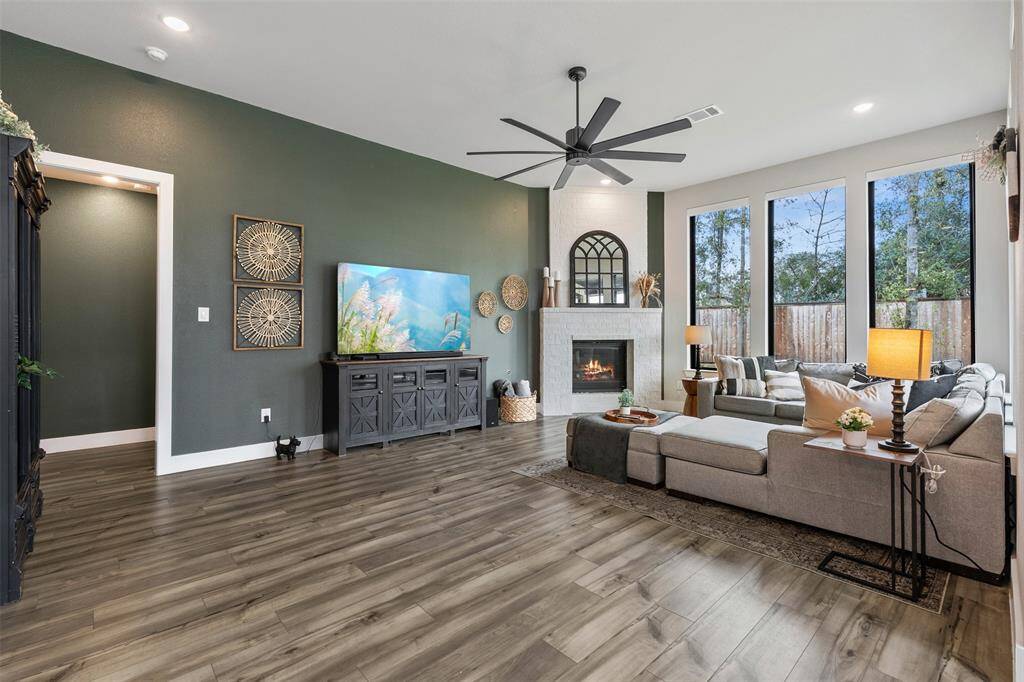 This spacious living room features sleek wood flooring, and a cozy fireplace. Large windows provide ample natural light, and a stylish ceiling fan complements the contemporary decor.