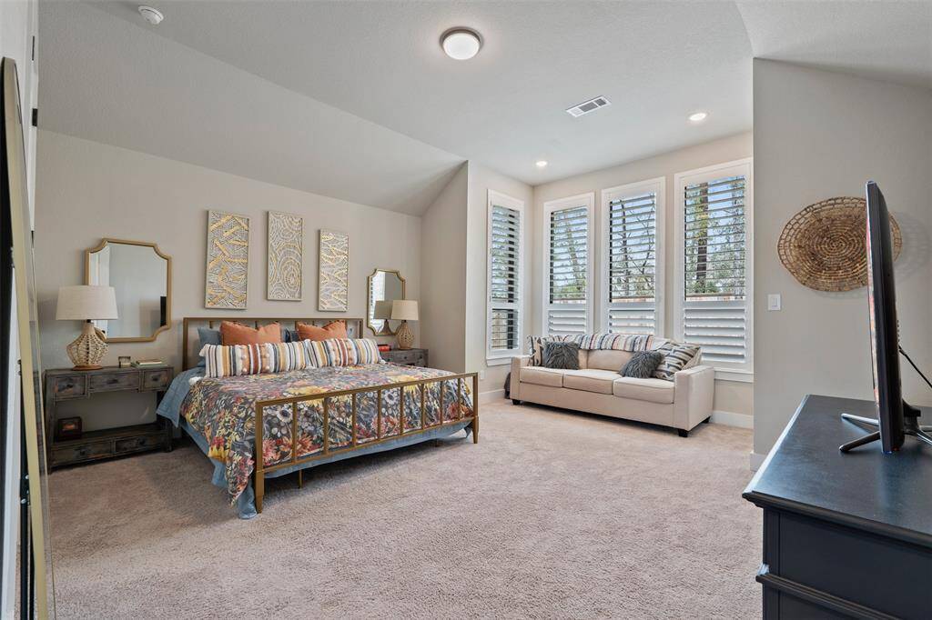 This spacious bedroom features ample natural light from multiple windows with shutters. There's a cozy sitting area large enough for a sofa, and the room has carpeted flooring for added comfort.
