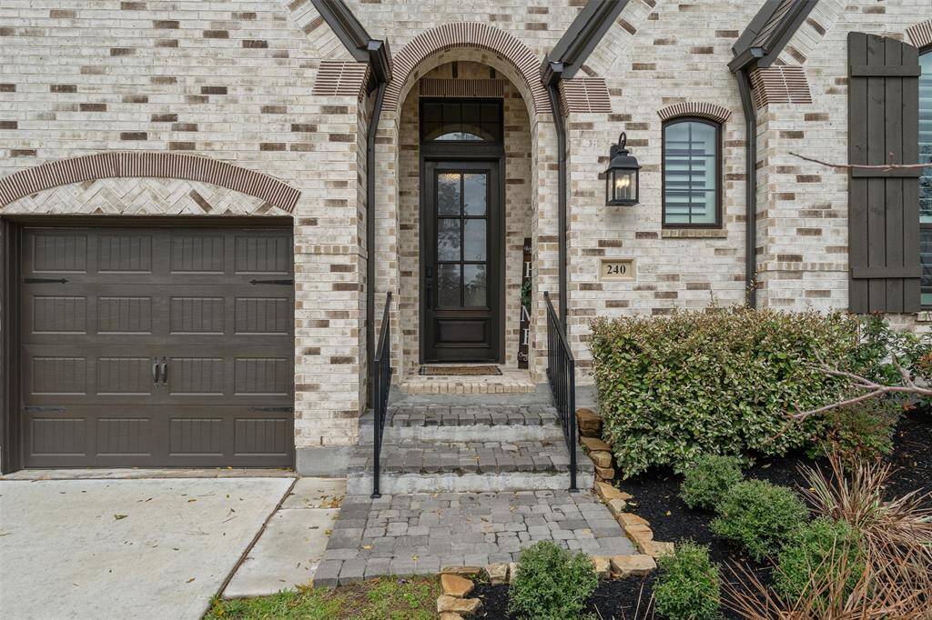 This Charming home entrance has a brick exterior. It features a dark, elegant front door with glass panes, a welcoming lantern-style light fixture. The entrance is framed by well-maintained shrubs and a stone pathway, adding to the curb appeal.