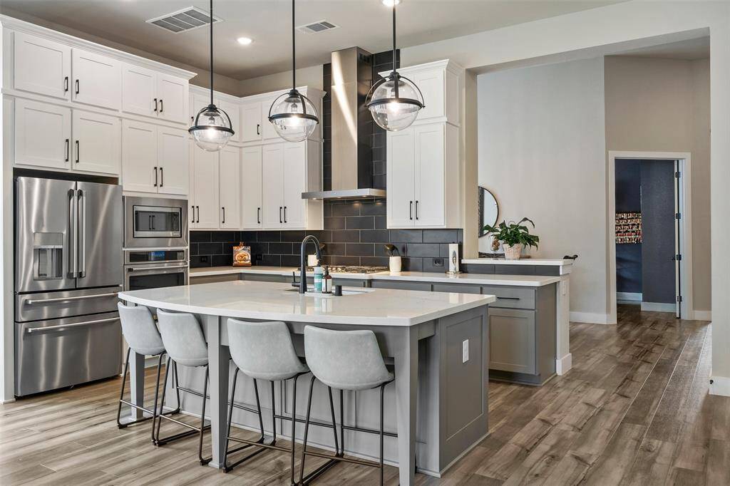 This modern kitchen, with sleek stainless steel appliances,  has a large island with seating, and contemporary pendant lighting. The cabinetry is a mix of white and gray tones, complemented by a stylish dark backsplash. The open layout and wood flooring enhance the spacious, inviting feel.