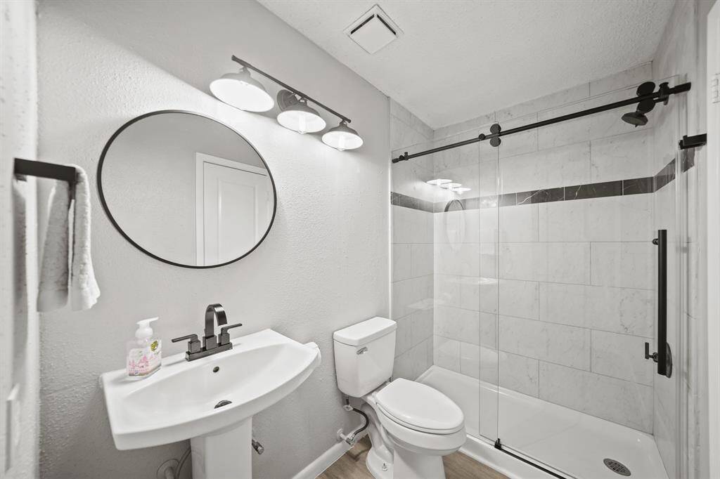 A private water closet has a second sink with framed mirror & wall mounted lighting above, a ceramic tile surrounded & glass enclosed walk-in shower & luxury vinyl plank hardwood flooring.