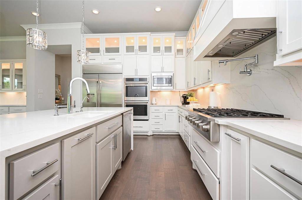 This kitchen features a modern design with sleek white cabinetry, premium stainless steel appliances, and a spacious island with a quartz countertop. It has ample storage, a large gas cooktop , and elegant pendants lighting, creating a bright and functional cooking space.