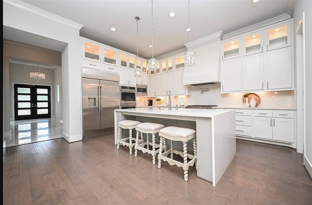 This modern kitchen with sleek white cabinetry, luxury stainless steel appliances, and a spacious island with a quartz countertop. waterfall. The room is well-lit with pendant chandeliers  and features a clean, minimalist design.