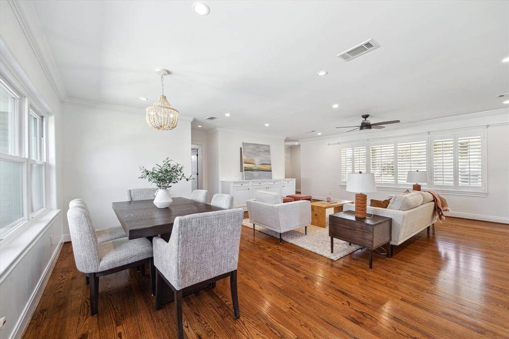 This open-plan living and dining area features hardwood floors, recessed lighting, custom built-in cabinets, custom crown molding, plantation shutters, ample natural light from large windows, and a neutral color palette.