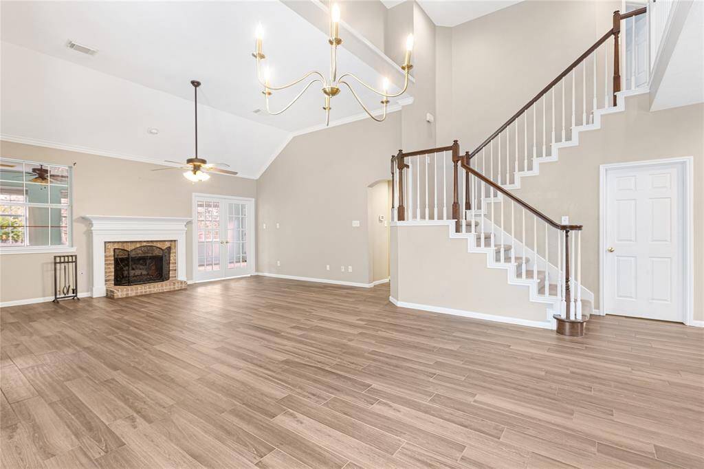 A spacious, well-lit living area with high ceilings, modern chandeliers, and a classic fireplace. It features a beautiful wooden staircase leading to the second level, elegant flooring, and large windows with a glass door providing outdoor access to a sunroom