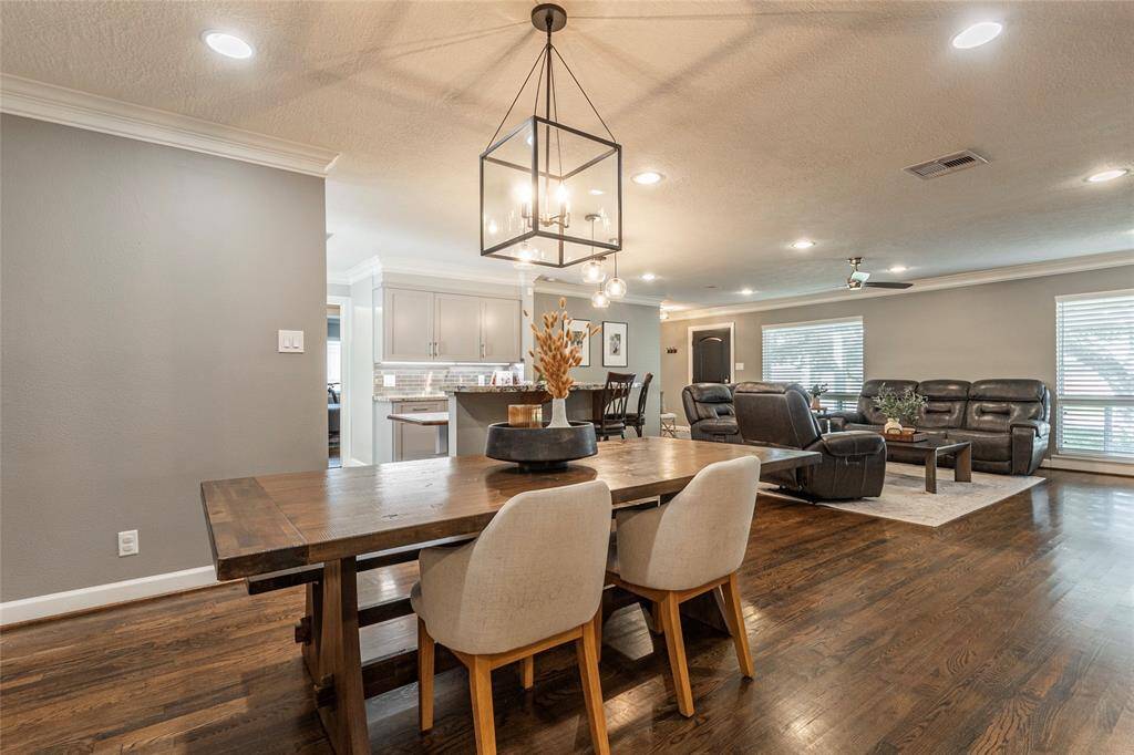 This open-concept living and dining area features hardwood floors, modern lighting, and neutral tones. The space is inviting with a large dining table, comfortable seating area, and ample natural light from the windows. The kitchen is visible in the background, offering a cohesive flow throughout the home.