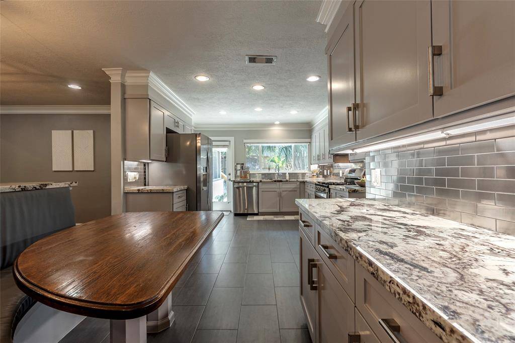 This kitchen features modern gray cabinetry, stainless steel appliances, and a sleek tile backsplash. The space is brightened by recessed lighting and a large window, with ample counter space and a cozy breakfast bar.