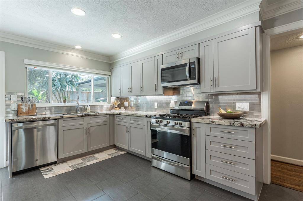 This modern kitchen features sleek gray cabinetry, stainless steel appliances, and granite countertops. A large window provides natural light, and the space is complemented by a stylish tile backsplash and recessed lighting.