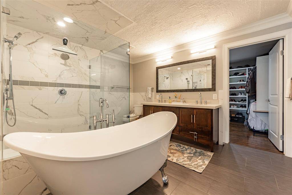 This elegant primary bathroom features a freestanding tub, heated floors, a spacious glass-enclosed shower with modern fixtures, and a large double vanity with ample storage. The room is well-lit with sleek lighting, and it connects to a walk-in closet, adding convenience and luxury.