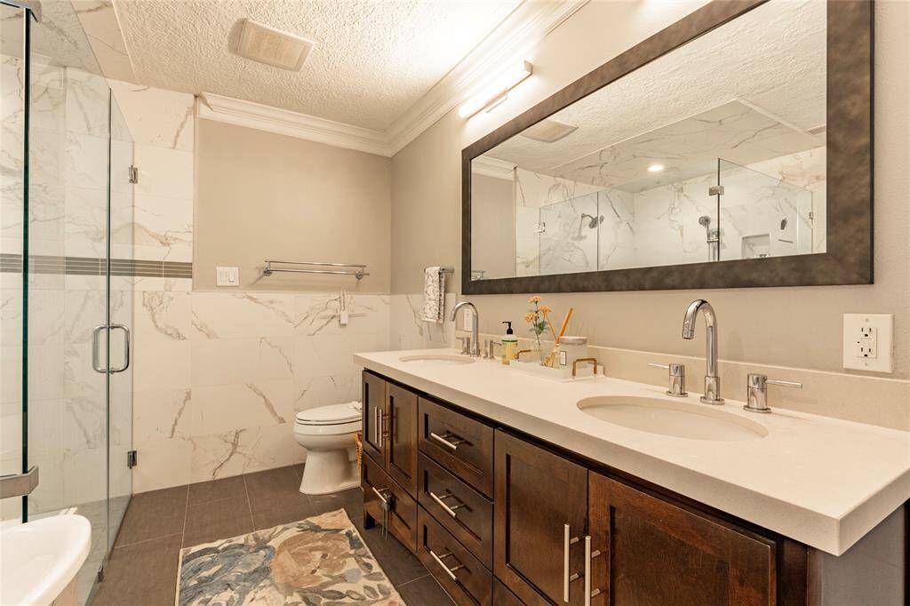 This remodeled primary bathroom features a modern design with a large double vanity, sleek faucets, and ample storage space. It has a spacious glass-enclosed shower with marble-style tiles and a neutral color palette, creating a clean and elegant look.