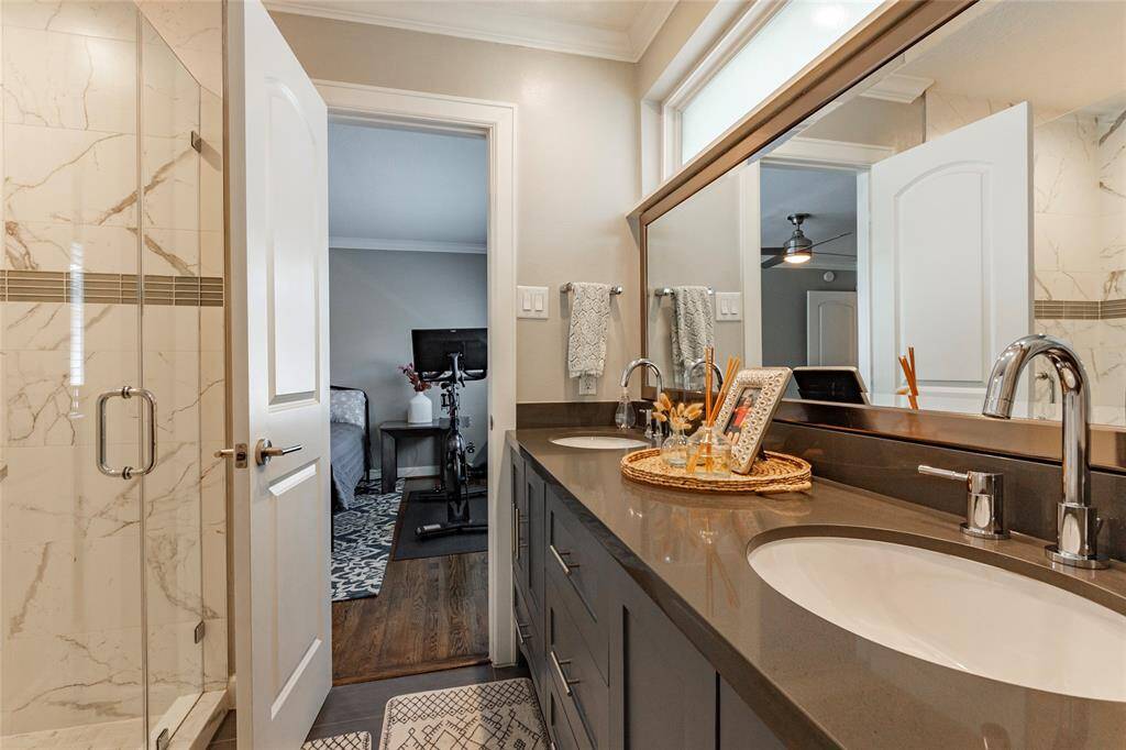 This stylish bathroom features a double vanity with sleek countertops and a large mirror. There is a modern, glass-enclosed shower with marble-style tiles. The room is well-lit and connects to a bedroom with hardwood flooring.