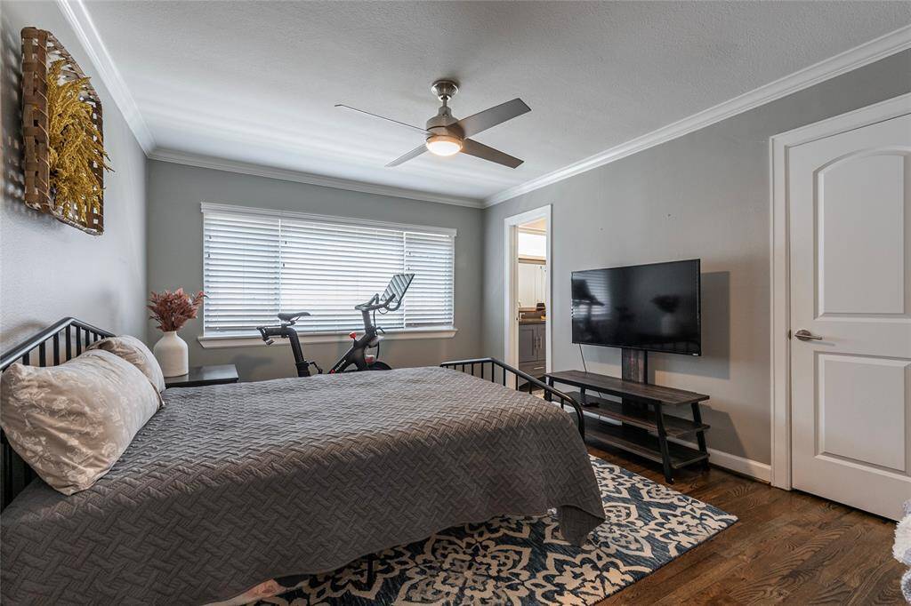 This bedroom features a cozy setup with a double bed, a large window for natural light, and a ceiling fan. It includes a TV on a stand, an exercise bike, and a door leading to an en-suite bathroom. The room has hardwood floors and neutral gray walls for a modern feel.