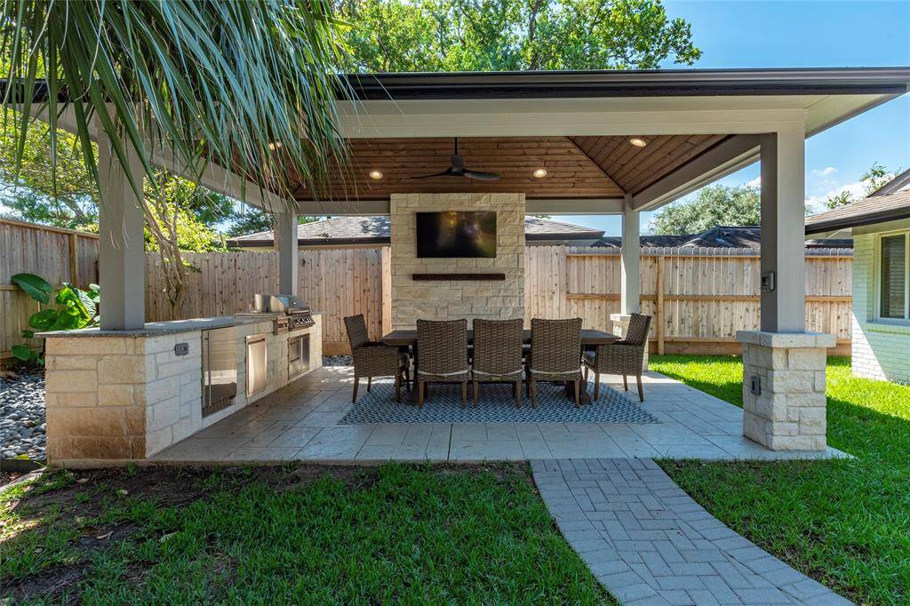 This is a stylish outdoor patio featuring a covered dining area with a ceiling fan and built-in grill. It includes a stone fireplace with a mounted TV, surrounded by a privacy fence and lush greenery, perfect for entertaining.