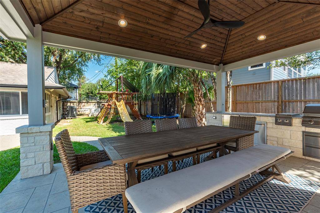 This inviting backyard features a covered patio with a wooden ceiling and recessed lighting, perfect for outdoor dining. It includes a large table with wicker chairs and a bench, set on a stylish rug. The space also boasts an outdoor kitchen with a built-in grill, and a view of the lush garden with a children's playset.