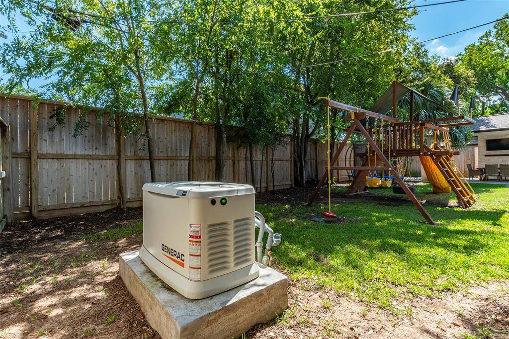 This backyard features a wooden playset with swings and a slide, perfect for children. A standby generator is installed on a concrete pad, providing reliable backup power. The yard is fenced and shaded by trees, offering privacy and a serene outdoor space.