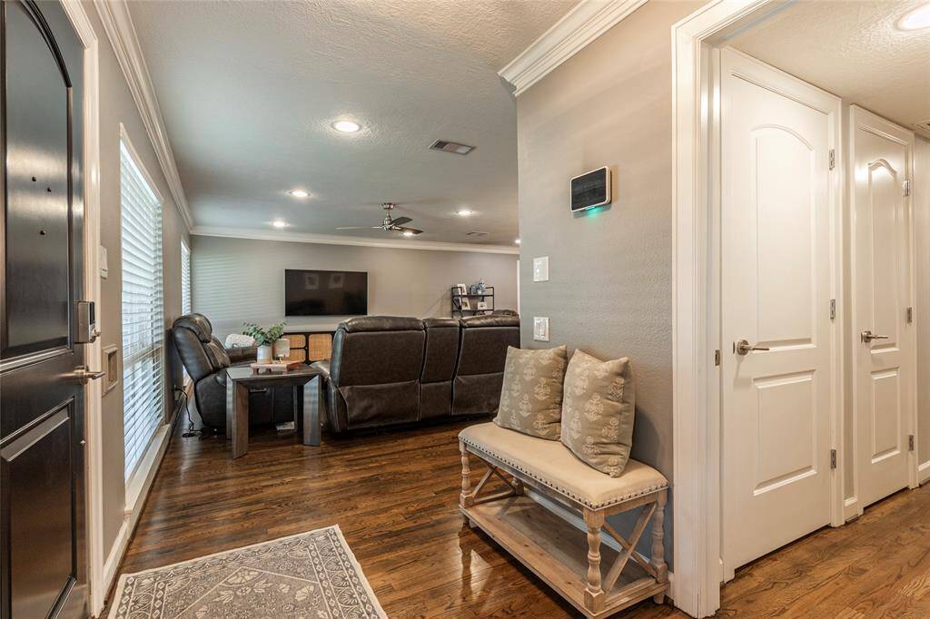 This inviting living room features polished hardwood floors, neutral walls, and ample natural light from large windows. It includes a cozy seating area with a large TV and an entryway with a decorative bench, creating a welcoming atmosphere.