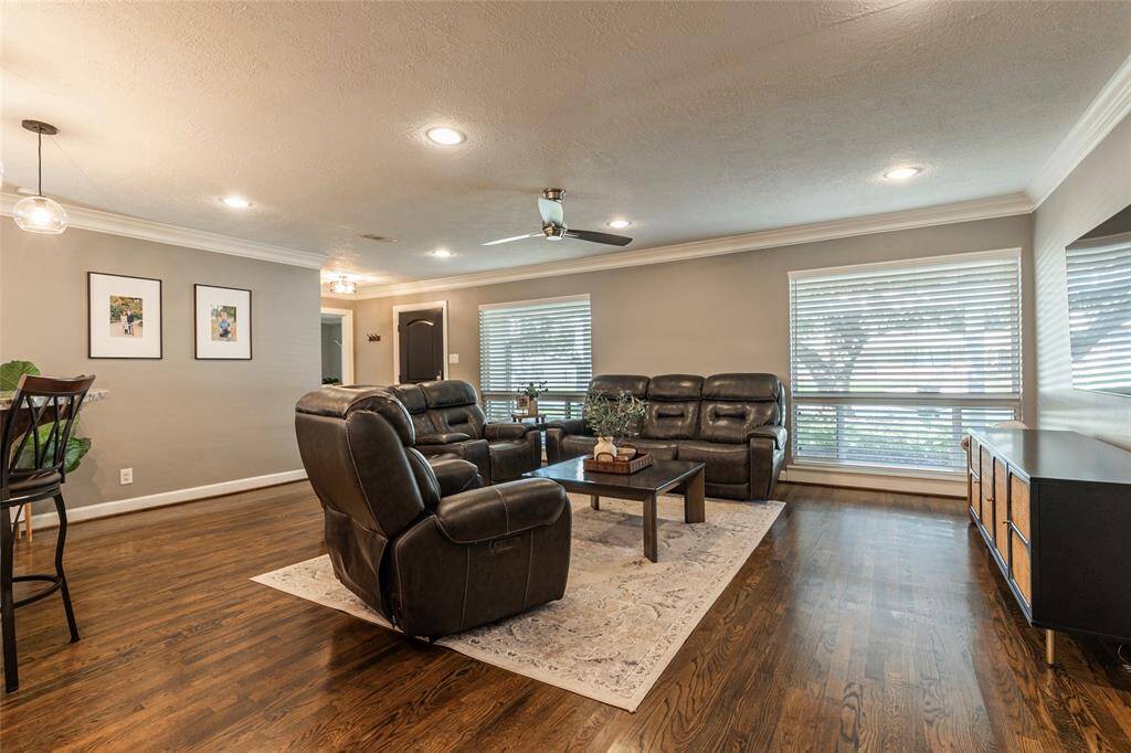 This spacious living room features hardwood floors, comfortable leather seating, and large windows for natural light. It includes modern ceiling lights, a ceiling fan creating a warm and inviting atmosphere.