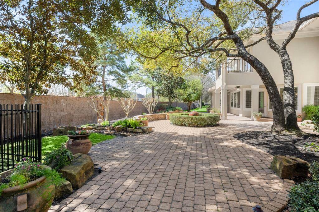 This inviting backyard features a spacious paver patio, mature trees providing ample shade, and well-maintained landscaping with potted plants and greenery. Perfect for outdoor relaxation and entertaining.