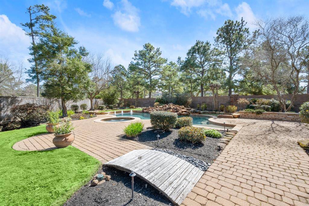 This backyard features a beautiful pool with a waterfall, surrounded by lush landscaping and mature trees. A stone patio provides ample space for outdoor entertaining, and a small wooden bridge adds charm to the garden area.
